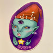 Art sticker of a green skinned elf with a purple third-eye on their forehead wearing a red cone hat with white mushrooms growing from underneath the hat. One hand is held up and the other is holding a magic wand with a sparkle coming from the tip. Under the head and between the two hands is a floating shining gold star. The background is purple.