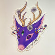 A purple deer-like creature with cream colored antlers and rainbow mushrooms growing from its head. Three daisies surround its antlers and it has a green third-eye in the middle of its forehead