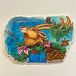 Art sticker of a deer’s top half appearing out of a patch of ferns and roses and lilies and shelf mushrooms. A waterfall flows out of the patch of greenery and there are three lily pads around the bottom. The deer’s antlers are covered in moss and algae and two butterflies are landing on it. A green dragonfly zooming around the front of the deer. 