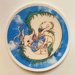 Circle sticker of a fluffy cream colored dragon with brown speckles and green hair down it’s back curled over itself slightly while flying on a blue sky background with white clouds