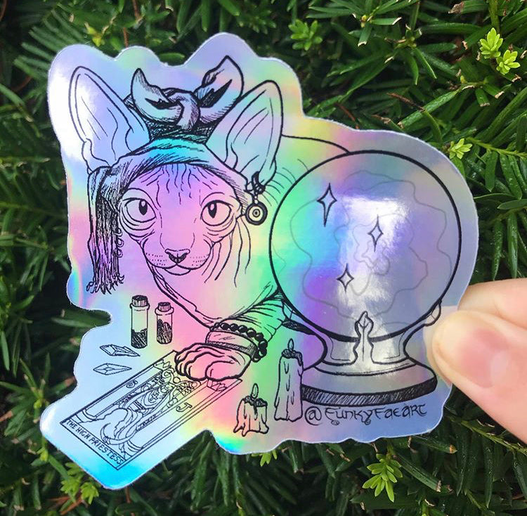 Rainbow holographic art sticker of a hairless cat with a headwrap hunched over a tarot card of The High Priestess surrounded in crystals and melting candles. Held in front of a green plant outside.