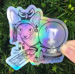 Load image into Gallery viewer, Rainbow holographic art sticker of a hairless cat with a headwrap hunched over a tarot card of The High Priestess surrounded in crystals and melting candles. Held in front of a green plant outside.
