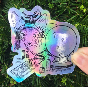 Rainbow holographic art sticker of a hairless cat with a headwrap hunched over a tarot card of The High Priestess surrounded in crystals and melting candles. Held in front of a green plant outside.